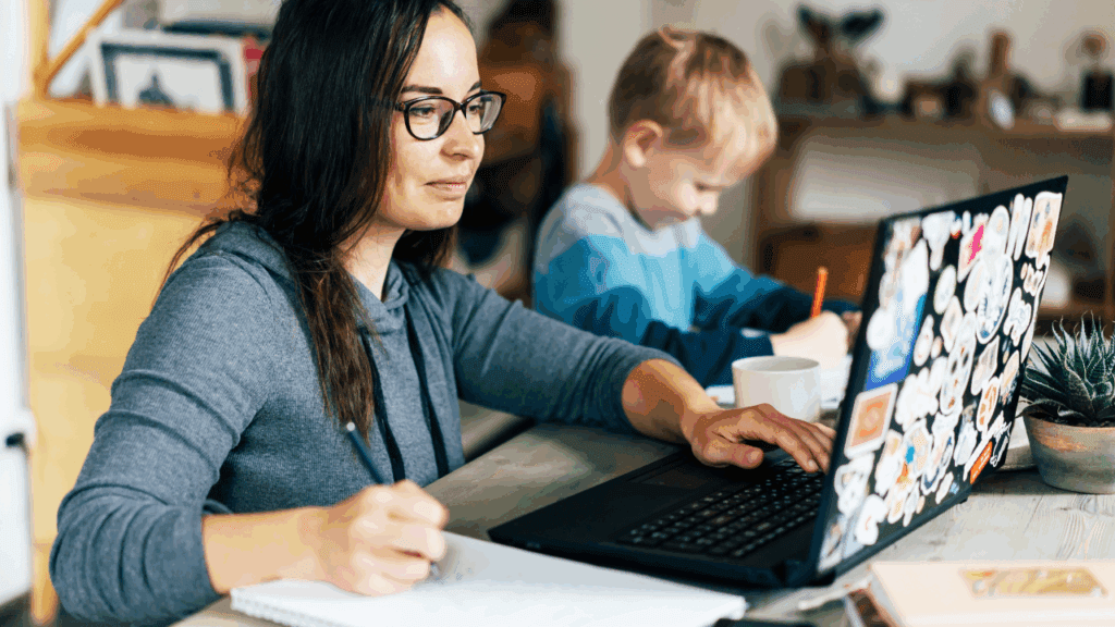 woman wearing glasses and a child sit together at a table with a laptop, embodying the struggle of working mom guilt.